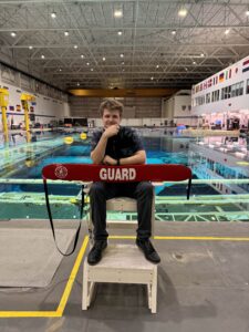 Student sitting and holding lifeguard floaty in front of pool with space craft pieces submerged (Neutral Buoyancy Lab JSC). 